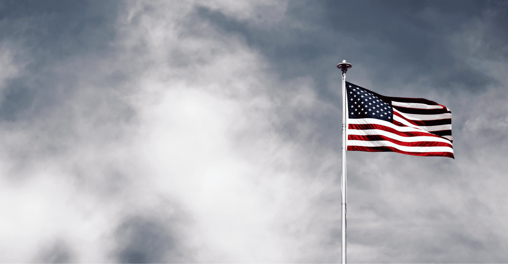 Flaga USA na tle zachmurzonego szarawo-niebieskiego nieba.