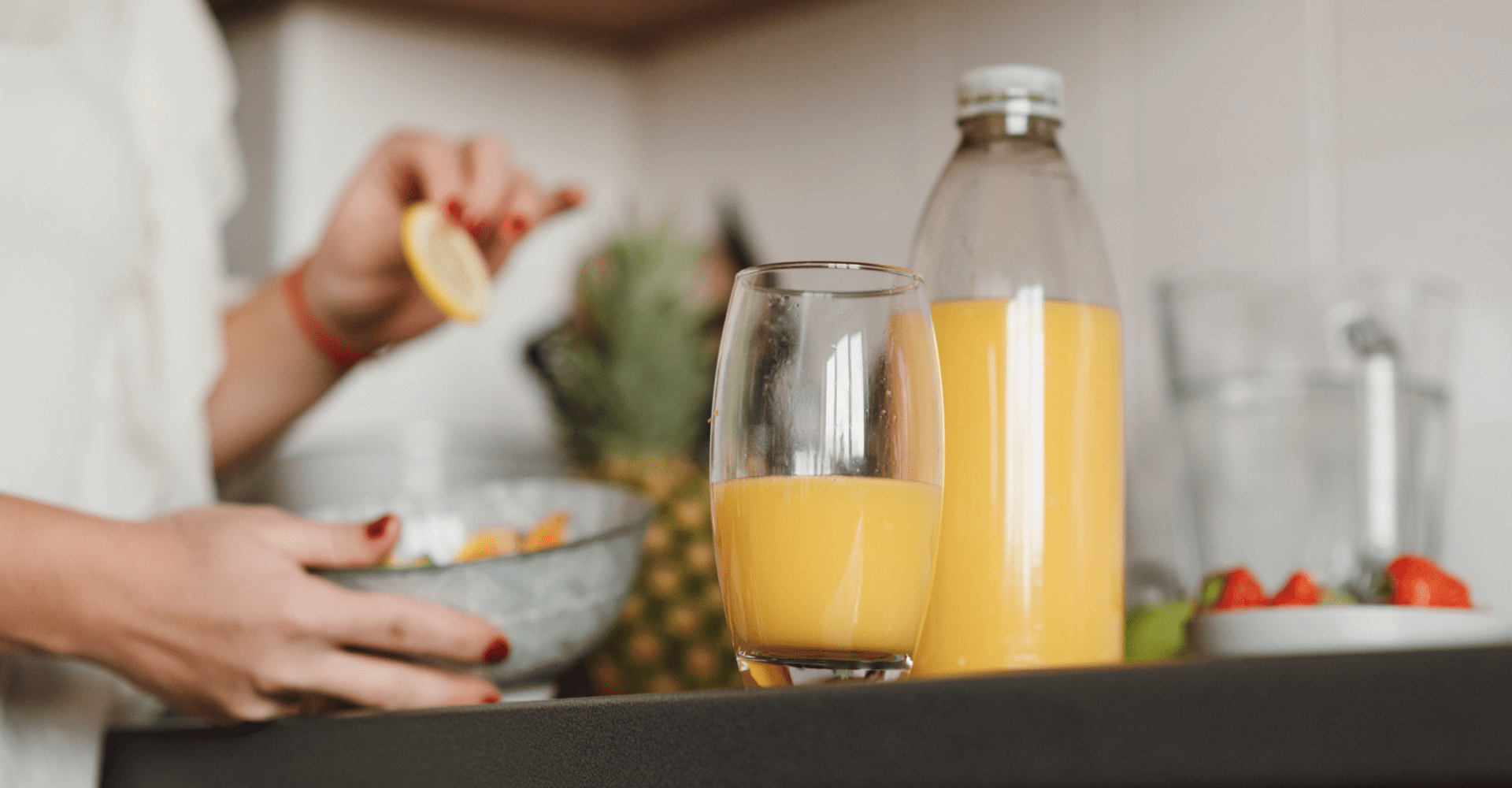 Kuchnia, na blacie stoi butelka i szklanka ze smoothie w kolorze żółtym. Z tyłu stoi osoba, trzyma miskę w jednej ręce, a w drugiej cytrynę