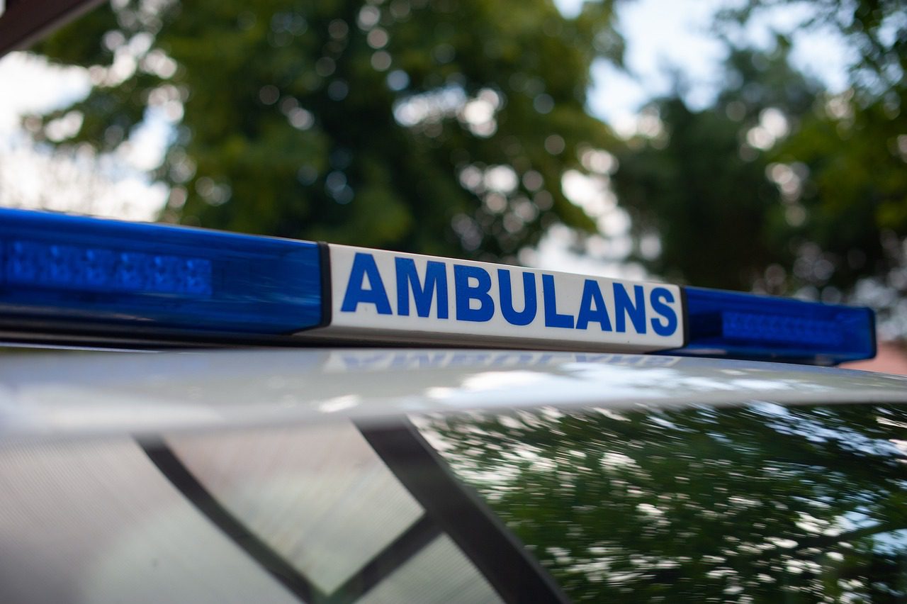 Niebieskie światła z napisem ambulans na dachu karetki pogotowia. W tle drzewa.