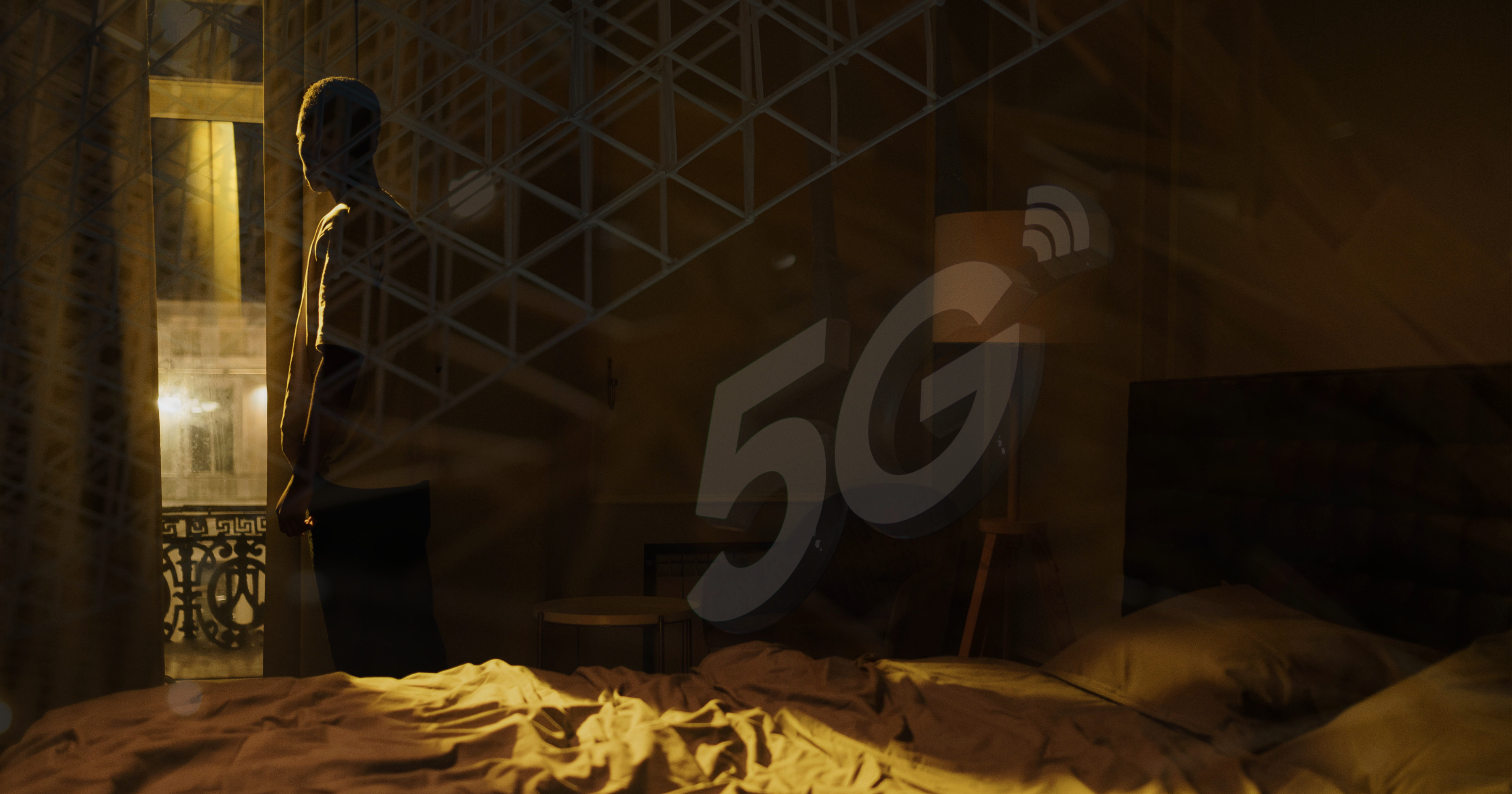 Mężczyzna stoi przy oknie, na zewnątrz jest ciemno. Na zdjęciu przebija się symbol 5G