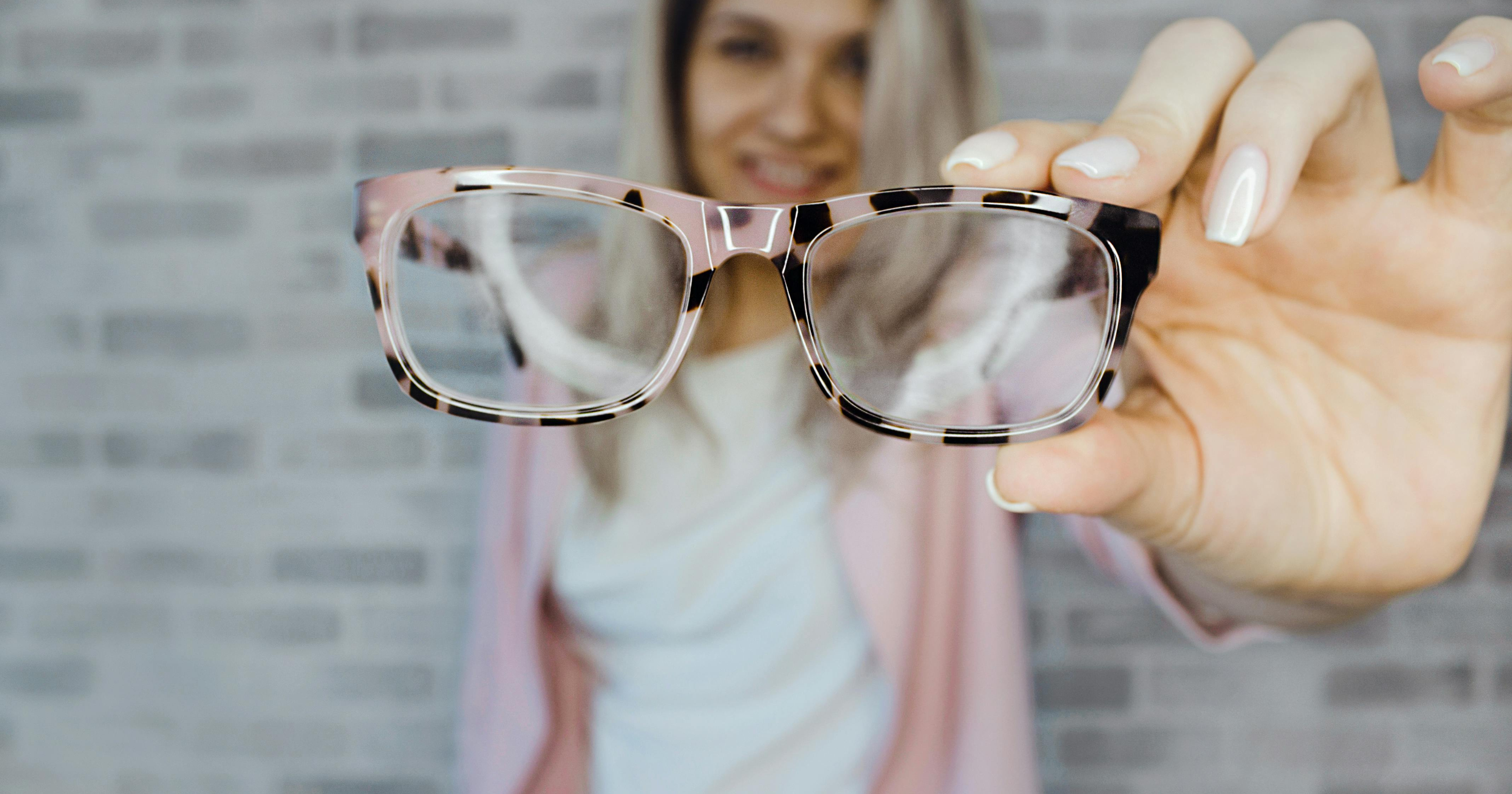 Fokus na okulary w prążki trzymane przez kobietę blondynkę ubraną w białą koszulkę i jasno-różowy sweter