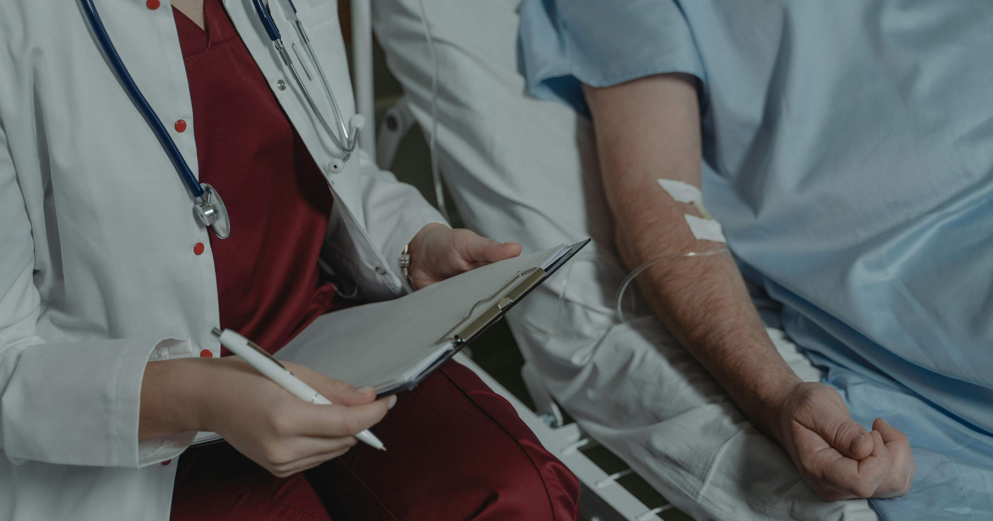 Lekarka ubrana w kitel i z lekarskimi słuchawkami na szyi trzyma w ręce podkładkę z kartką i długopis. Siedzi na krześle przy łóżku szpitalnym, na którym leży osoba z wenflonem w ręku.