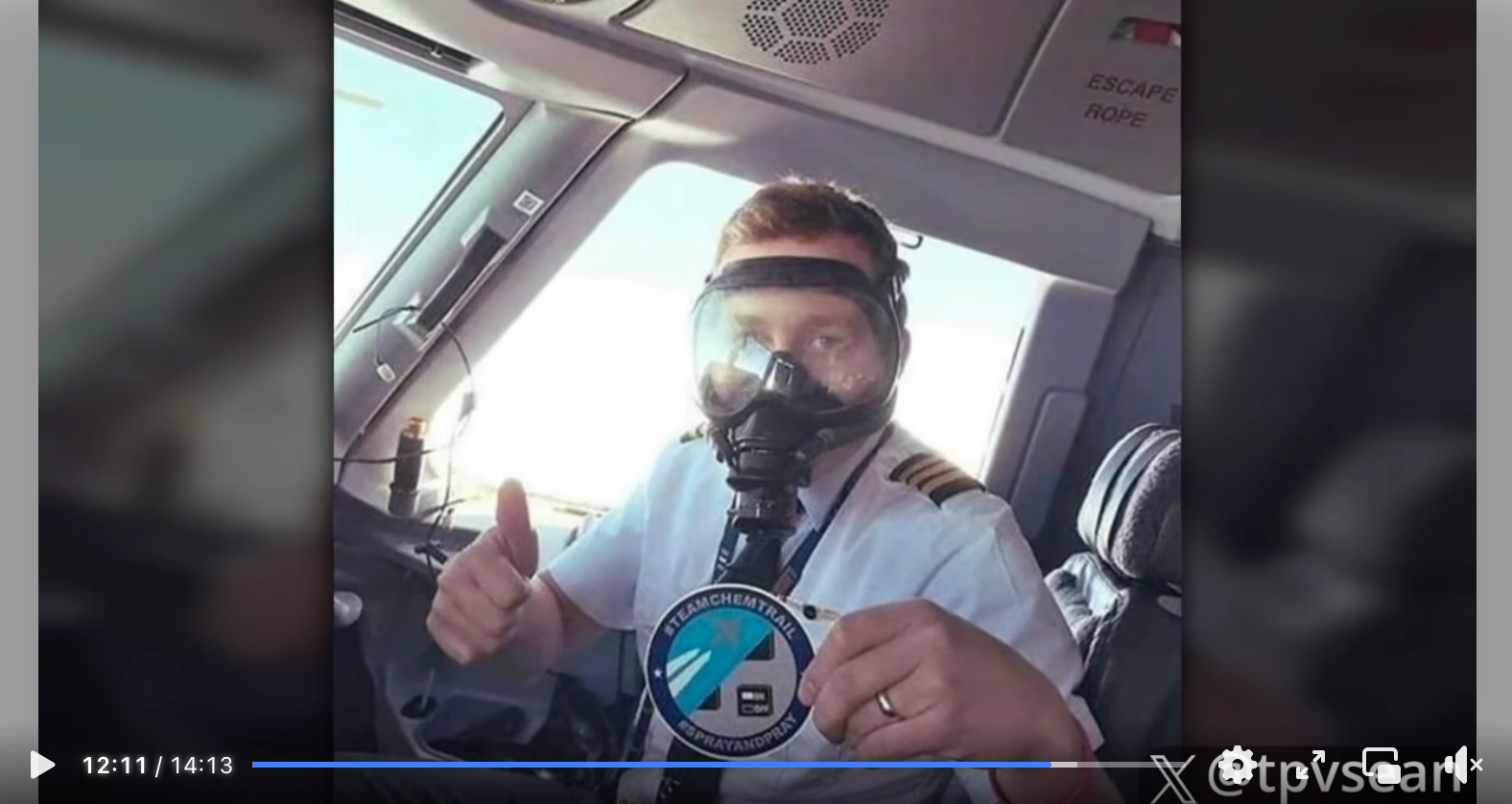 Kadr z omawianego filmu. Zdjęcie pilota w kokpicie, ubranego w maskę, trzymającego w ręku plakietkę z napisem TEAM CHEMTRAILS.